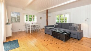 A spacious room with a wooden floor, a gray sofa, a dining table with chairs, and large windows. - Cottage life in intimate wooden cabin (Dronningmølle)