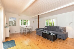 Um quarto amplo com piso de madeira, um sofá cinza, uma mesa de jantar com cadeiras e grandes janelas.