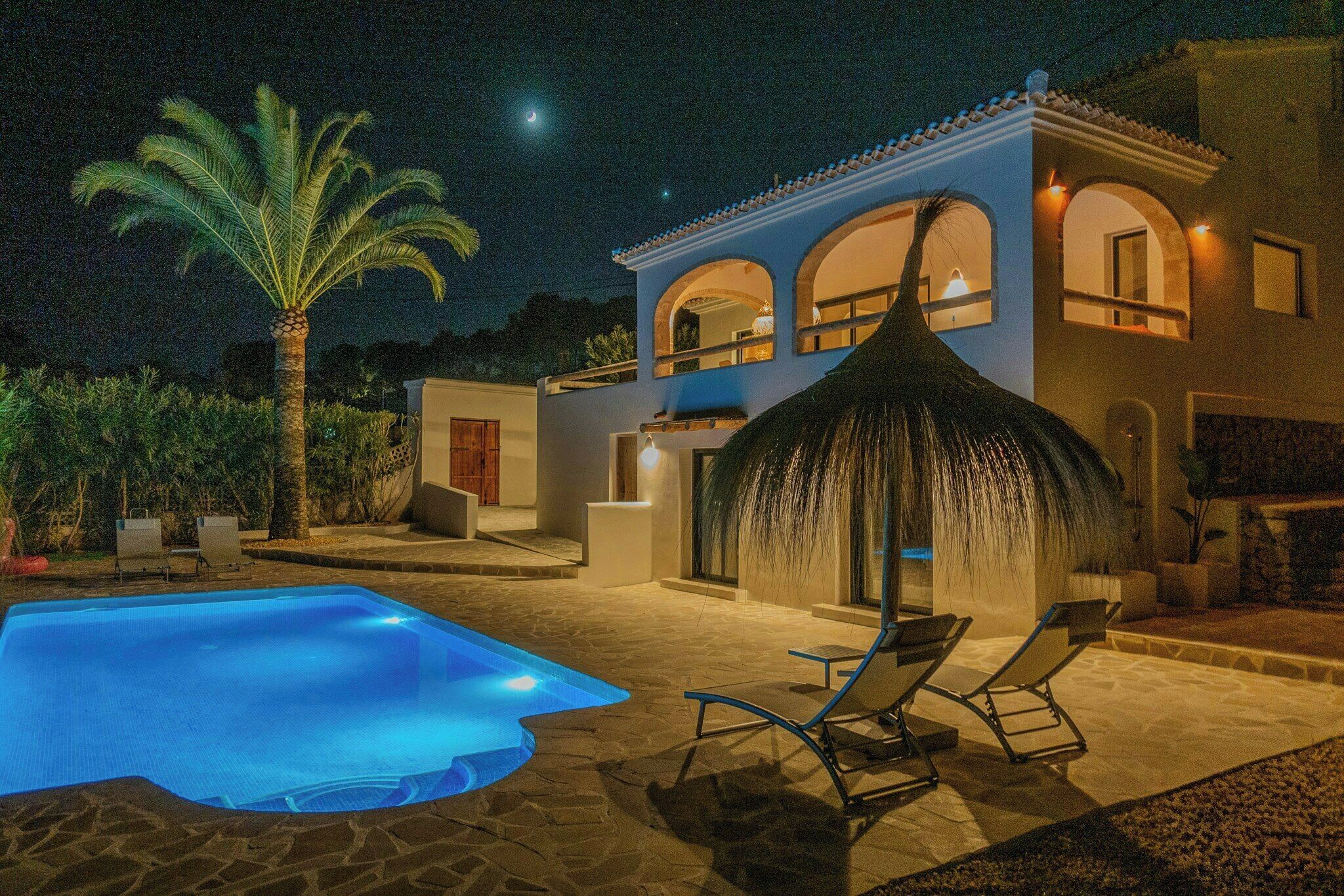 A poolside area with a pool, lounge chairs, and a house with arched windows and a thatched roof.
