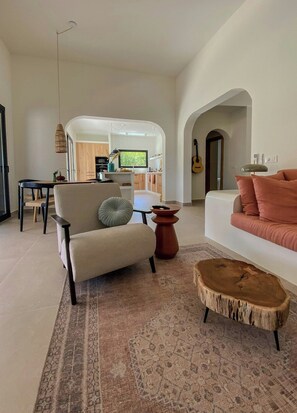 Ein modernes Wohnzimmer mit Sofa, Sessel und Couchtisch. Im Hintergrund ist eine Küche erkennbar.