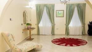 A room with a chair, a small table with a vase, a red rug, and two windows with green curtains.