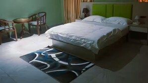 A bedroom with a bed, a desk, chairs, a ceiling fan, curtains, and a painting on the wall.