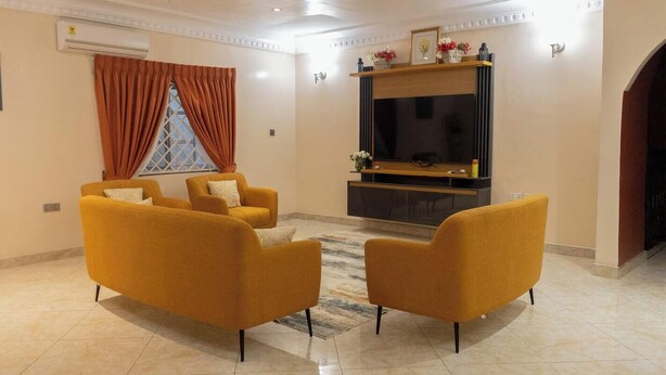 A living room with a flat-screen TV, two yellow armchairs, a window with orange curtains, and a fireplace. - Modern 3BR Luxury Diamond Hills Residency (Kumasi)