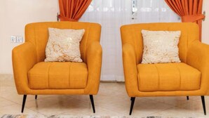 Two orange armchairs with matching cushions, positioned in front of a white curtain and a wall socket. - Modern 3BR Luxury Diamond Hills Residency (Kumasi)