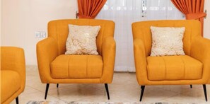 Deux fauteuils orange assortis de coussins correspondants, placés devant un rideau blanc et une prise de courant murale.