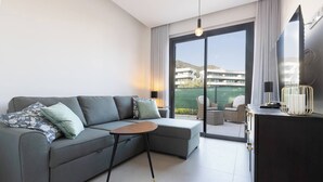 A modern living room with a grey sectional sofa, a small round table, and a dark wooden cabinet with a TV and decorative items. - Exclusive Garden Suite in Higueron (Fuengirola)