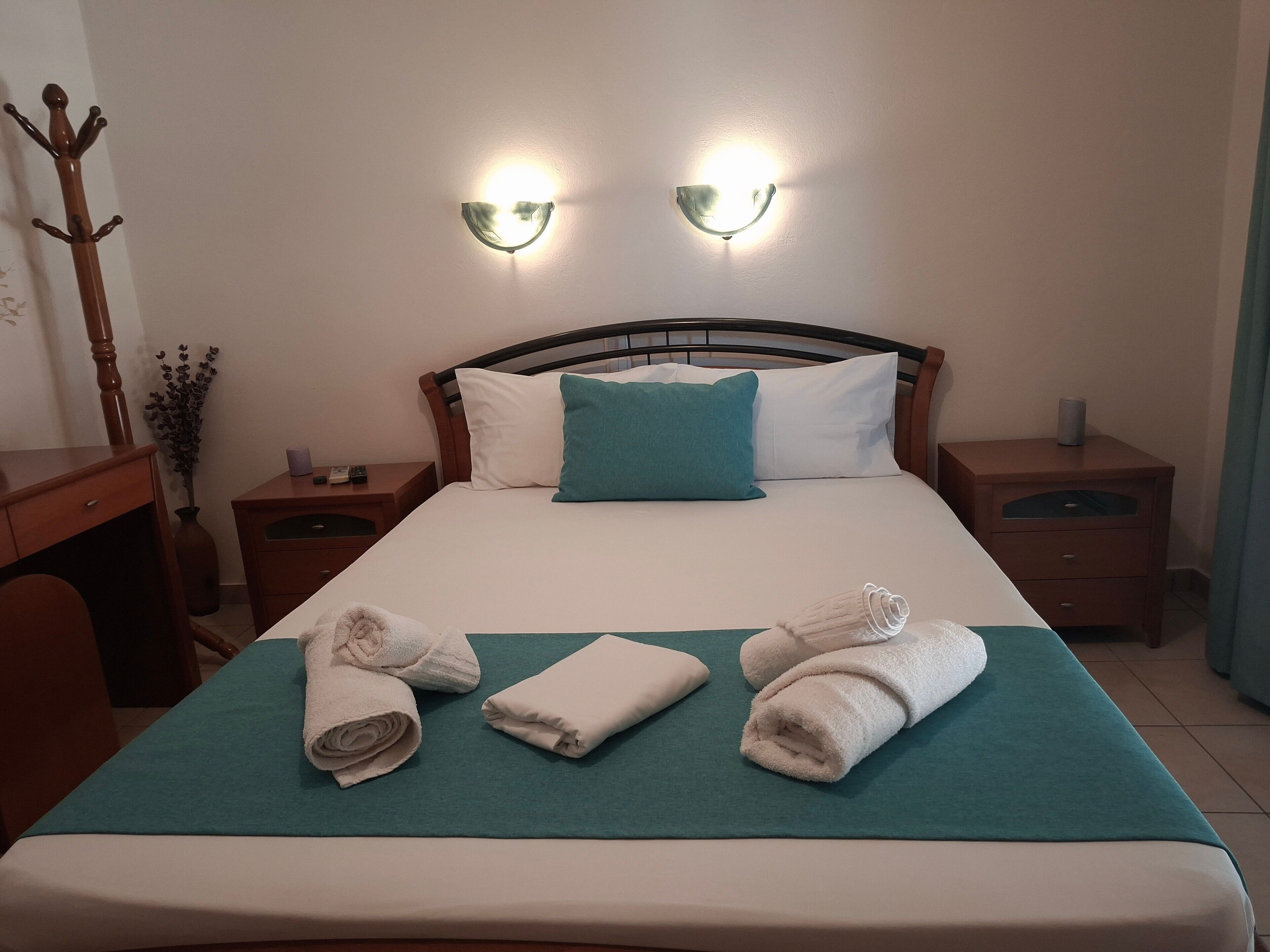 A neatly made bed with a teal bedspread, two white pillows, and three rolled towels. There are two bedside tables with lamps, a wooden coat rack, and a vase with dried flowers.