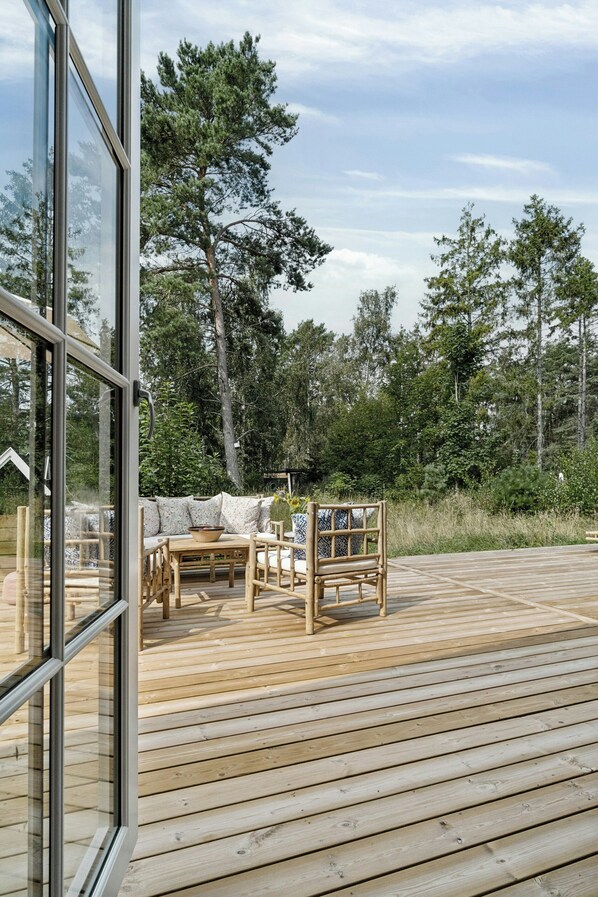 Un terrazzo in legno con zona salotto, parete vetrata e vista sugli alberi.