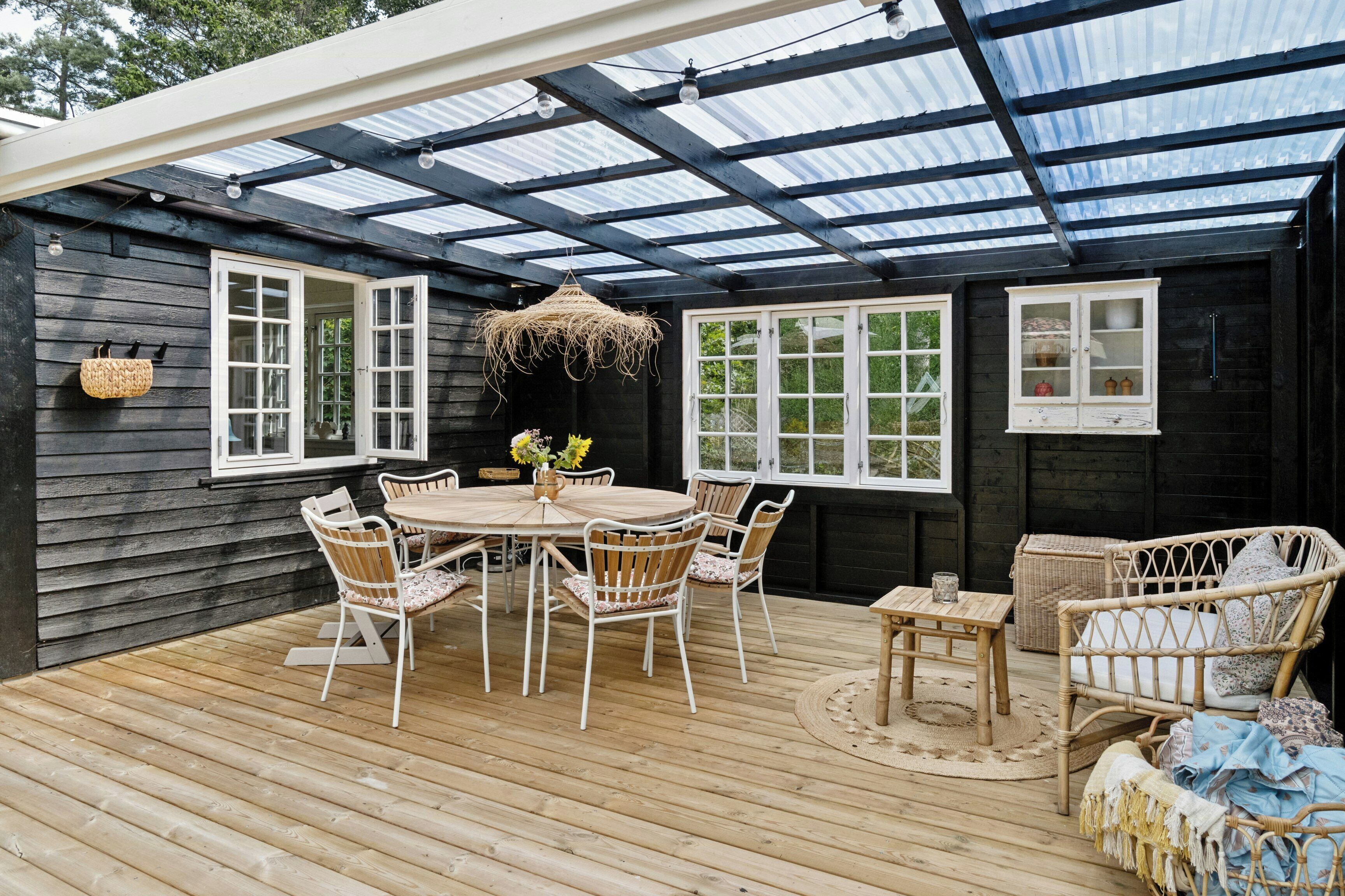 Eine überdachte Terrasse mit einem Holztisch und -stühlen, einem Korbsessel und einer hängenden Leuchte.