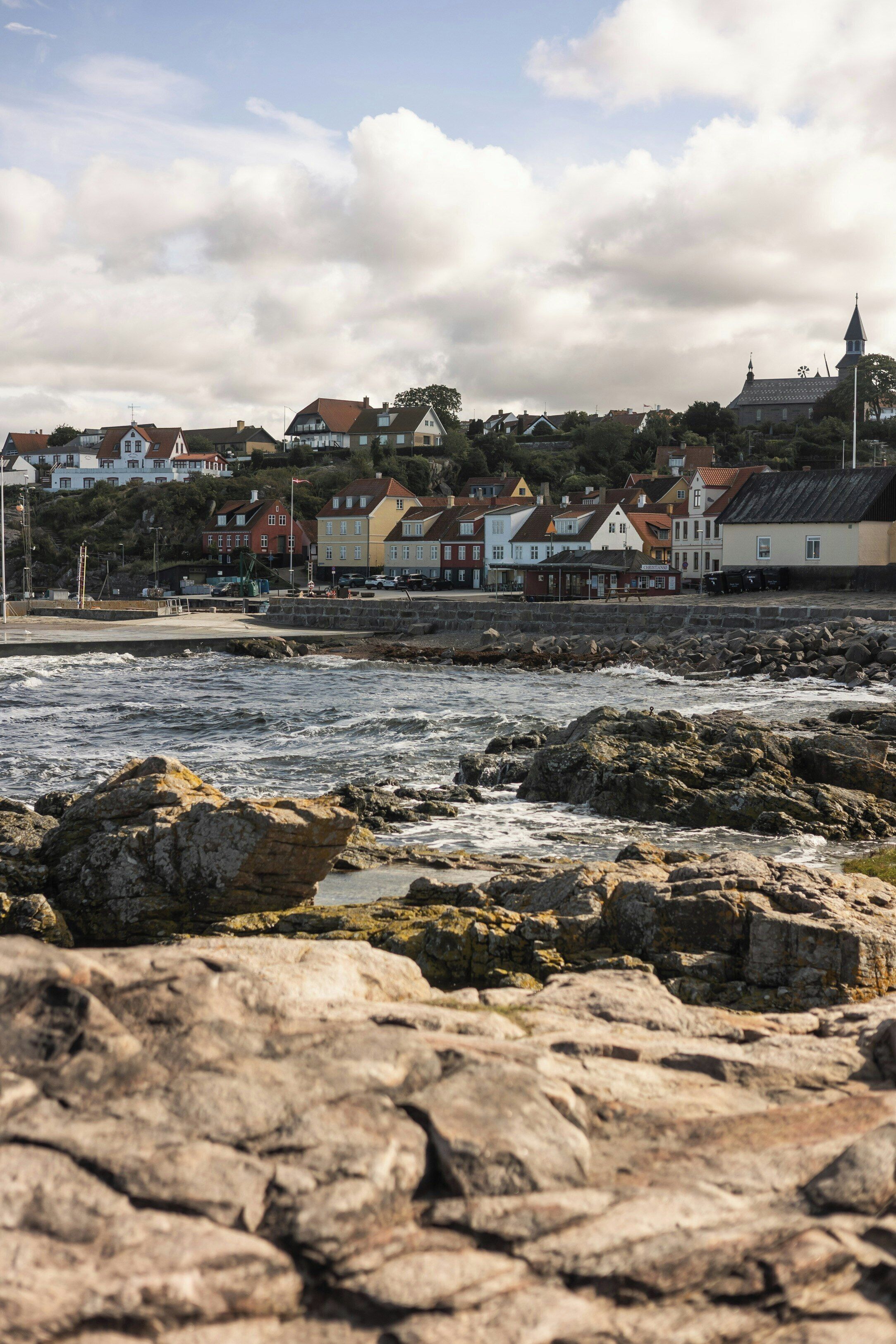 A coastal town with a rocky shoreline, houses with varied architecture, and a church steeple in the background.