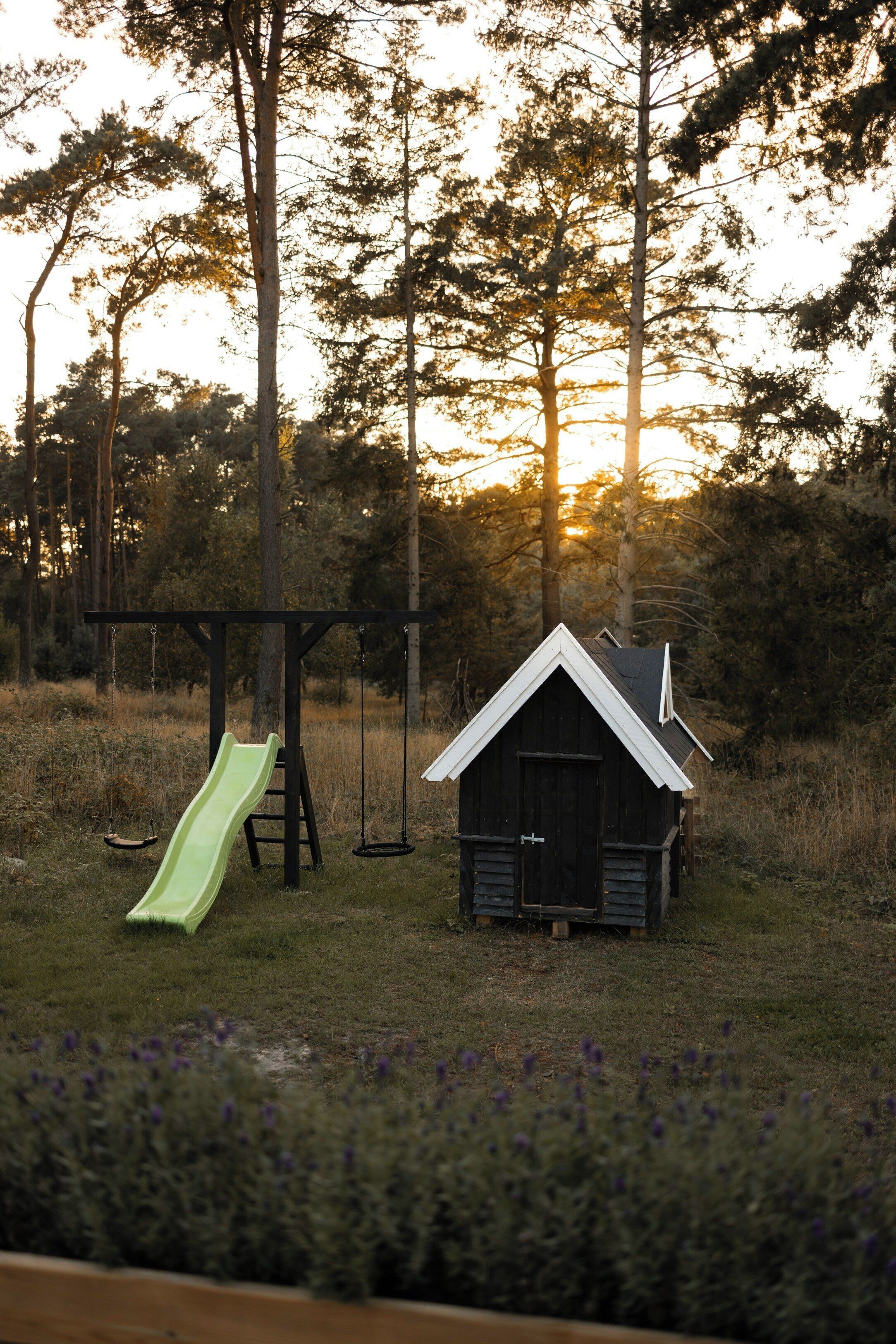 Une aire de jeux avec un toboggan et des balançoires, un petit abri et une structure ressemblant à une maison, le tout situé dans un espace verdoyant avec des arbres.