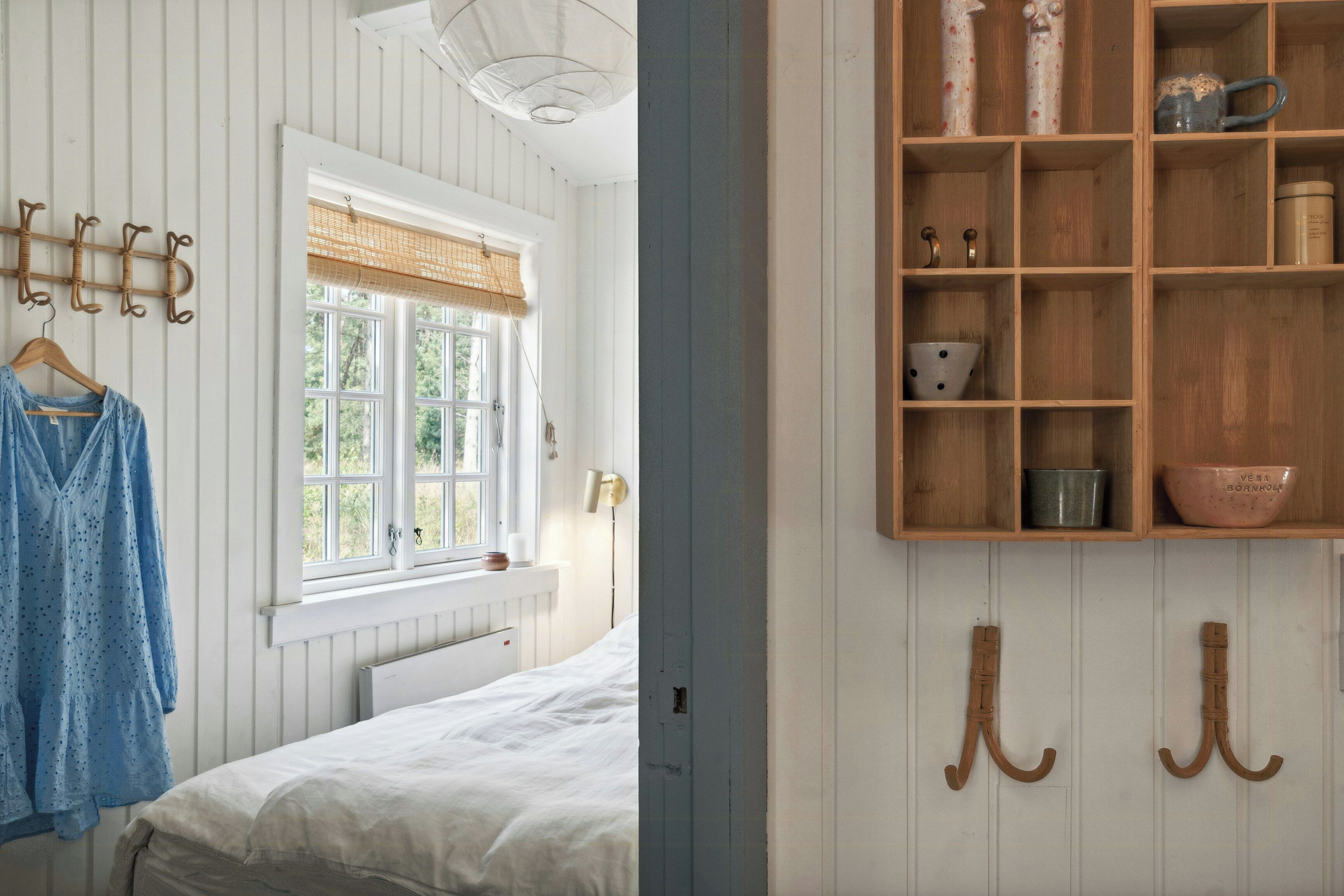 Un dormitorio con una cama, una ventana con vistas, un perchero y un estante de madera con varios objetos.
