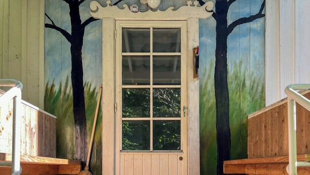 A wooden door with a window, a wooden bench, a broom, and a colorful rug on a wooden deck.