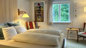 A hotel room with a bed, a chair, a table, a window, and a wall with framed pictures.