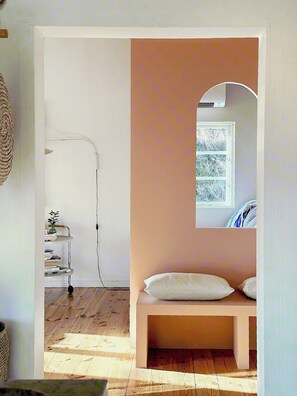 Ein Zimmer mit Holzboden, einer weißen Bank mit Kissen, einem kleinen Tisch mit einer Pflanze und einem Fenster mit Blick auf Bäume.