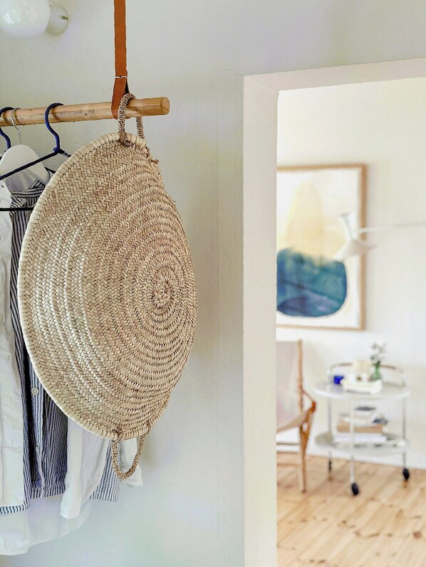 Una cesta tejida colgada en un perchero, una pared blanca y un vistazo a una habitación con una silla y una mesa.