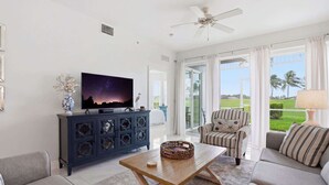 A living room with a TV, sofa, coffee table, and a view of a green landscape through large windows. - 514 - Golf Course View, First Floor, Sleeps 6 (Naples)