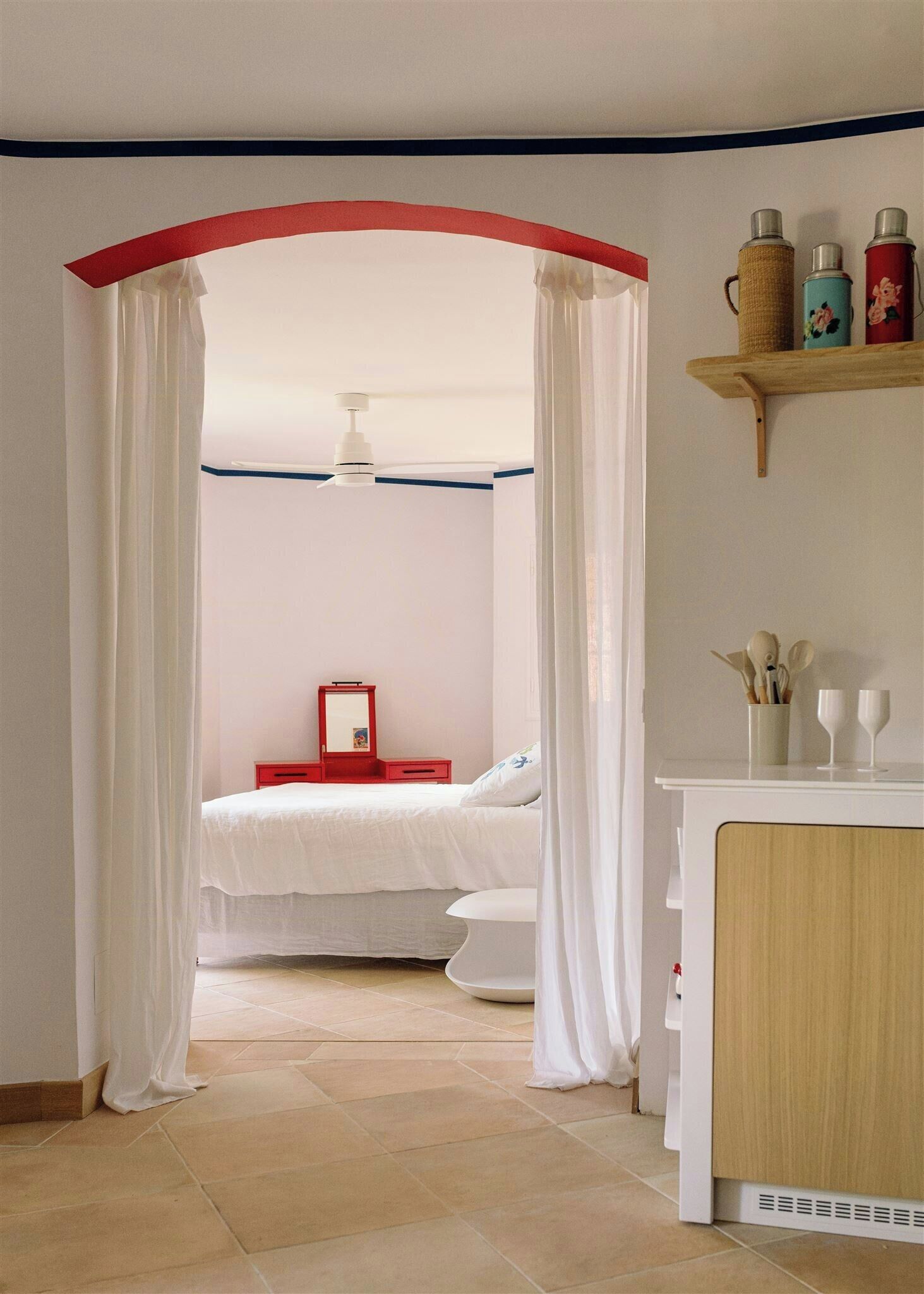 A bedroom with a bed, a red nightstand, a mirror, and a white bedside table. There is a wooden cabinet with a white countertop and a shelf above it.