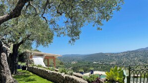A stone retaining wall, a grassy area, a stone building with a tiled roof, and a view of distant hills. - Exceptional apartment in Cannes' countryside (Spéracèdes)
