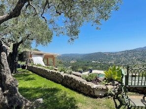 A stone retaining wall, a grassy area, a stone building with a tiled roof, and a view of distant hills. - Exceptional apartment in Cannes' countryside (Spéracèdes)