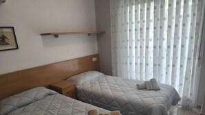 Habitació d'hotel amb dos llits, un capçal de fusta, una taula de nit i una finestra amb cortines translúcides.