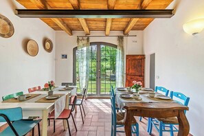 A dining area with wooden tables and chairs, a large window with curtains, and a view of greenery outside. - Early 1900s villa with pool near Ostra (Ostra)