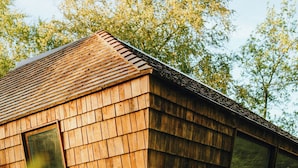 Une cabane en bois dotée d’un toit en pente, de grandes fenêtres et entourée d’un feuillage dense.