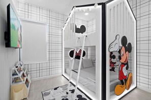 Un lit superposé pour enfants décoré aux couleurs de Mickey Mouse, une télévision et une bibliothèque dans une chambre moderne aux murs ornés d’un motif à carreaux.