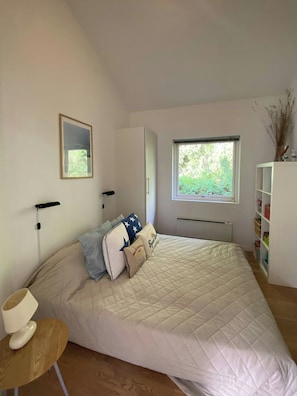 Un dormitorio con una cama grande, una mesita de noche, una ventana con vistas a zonas verdes, un espejo en la pared y un armario blanco.