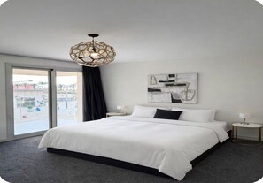 Ein Hotelzimmer mit einem großen Bett, einer geometrischen Deckenleuchte, Blick durch eine Schiebetür und einem an der Wand befestigten abstrakten Kunstwerk.