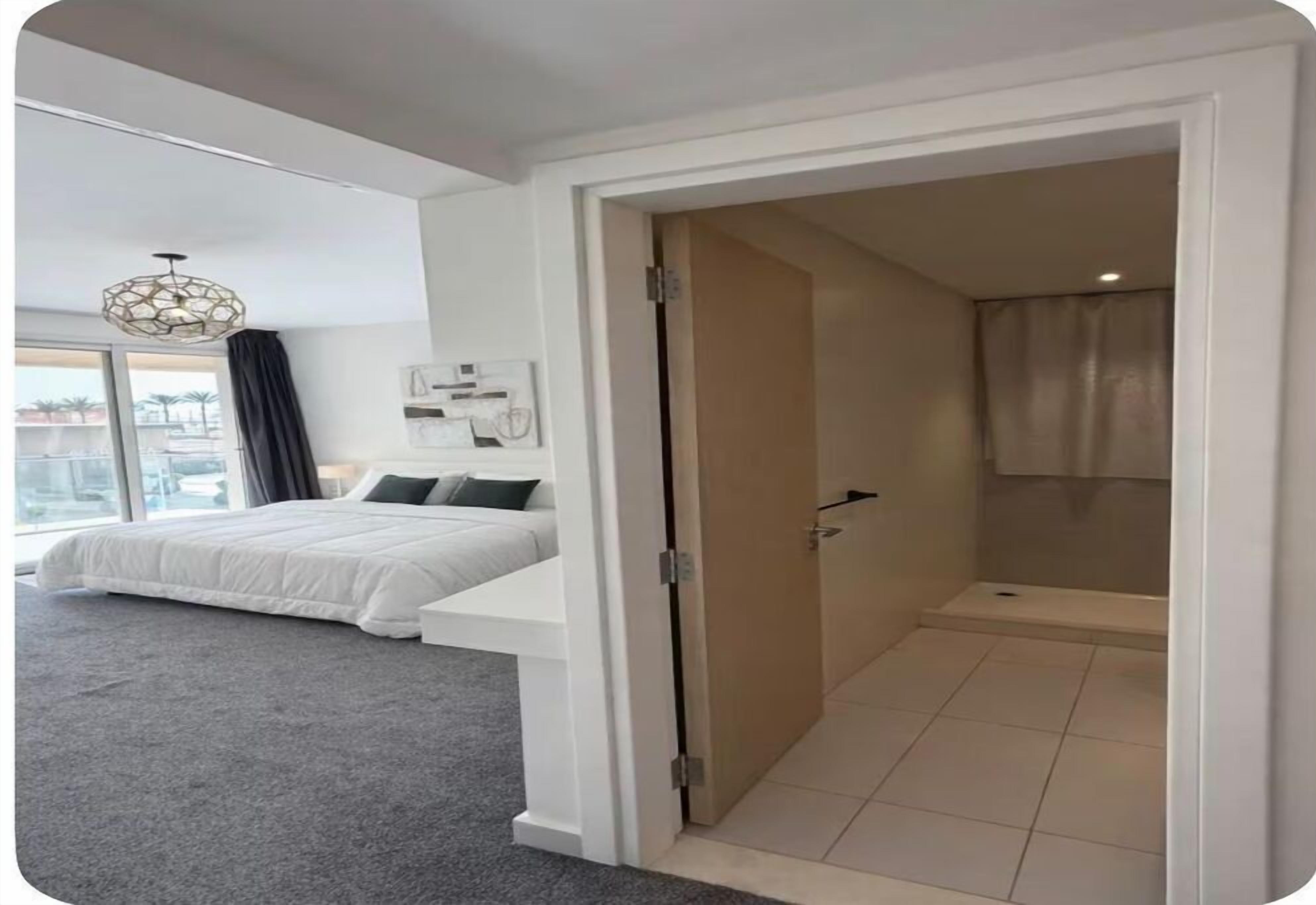 Une chambre à coucher comprenant un lit, une table de chevet, une fenêtre avec des rideaux, une porte donnant sur une salle de bain et un espace douche.