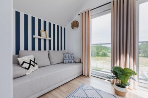 Un soggiorno moderno con un divano grigio, una parete a strisce blu e bianche, un'ampia finestra con tende e vista sul verde circostante.