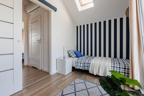 Un dormitorio con techo inclinado, cama con ropa de cama a cuadros, mesita de noche, planta pequeña y alfombra estampada.