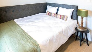 Ein Bett mit grauem Kopfteil, weißer Bettwäsche und einem gestreiften Kopfkissen. Neben dem Bett steht ein kleiner Tisch mit einer Lampe.