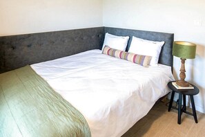 A bed with a grey headboard, white bedding, and a striped pillow. There is a small table with a lamp beside the bed.