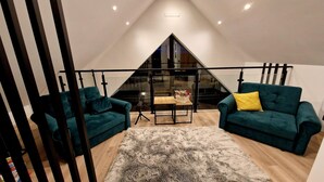 Une pièce de style loft avec un plafond en pente, deux canapés verts ornés de coussins jaunes, une table en verre avec un vase et un tapis moelleux.
