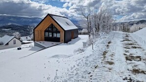 Un chalet moderne avec une façade en bois et de grandes fenêtres, entouré d’un paysage enneigé et de montagnes.