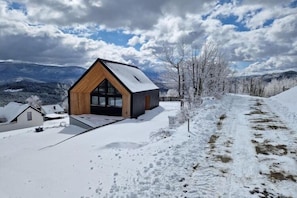 Una cabaña moderna con fachada de madera y grandes ventanales, rodeada por un paisaje nevado y montañas.