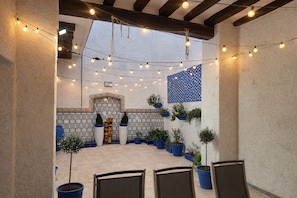 Un patio al aire libre con luces colgantes, plantas en macetas y un muro de azulejos con un diseño decorativo.