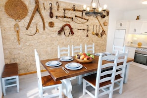 Un comedor con pared de piedra, mesa de madera, sillas blancas y un cuenco de frutas.