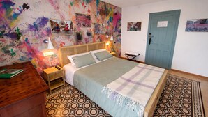 A bedroom with a bed, nightstands, a wall with colorful artwork, a door, and patterned rugs.