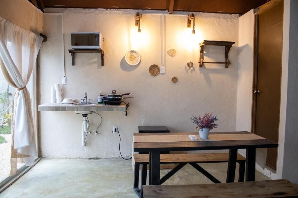 Una kitchenette pequeña con microondas, pileta y cocina, una mesa de madera con planta en maceta y un ventanal con cortinas.