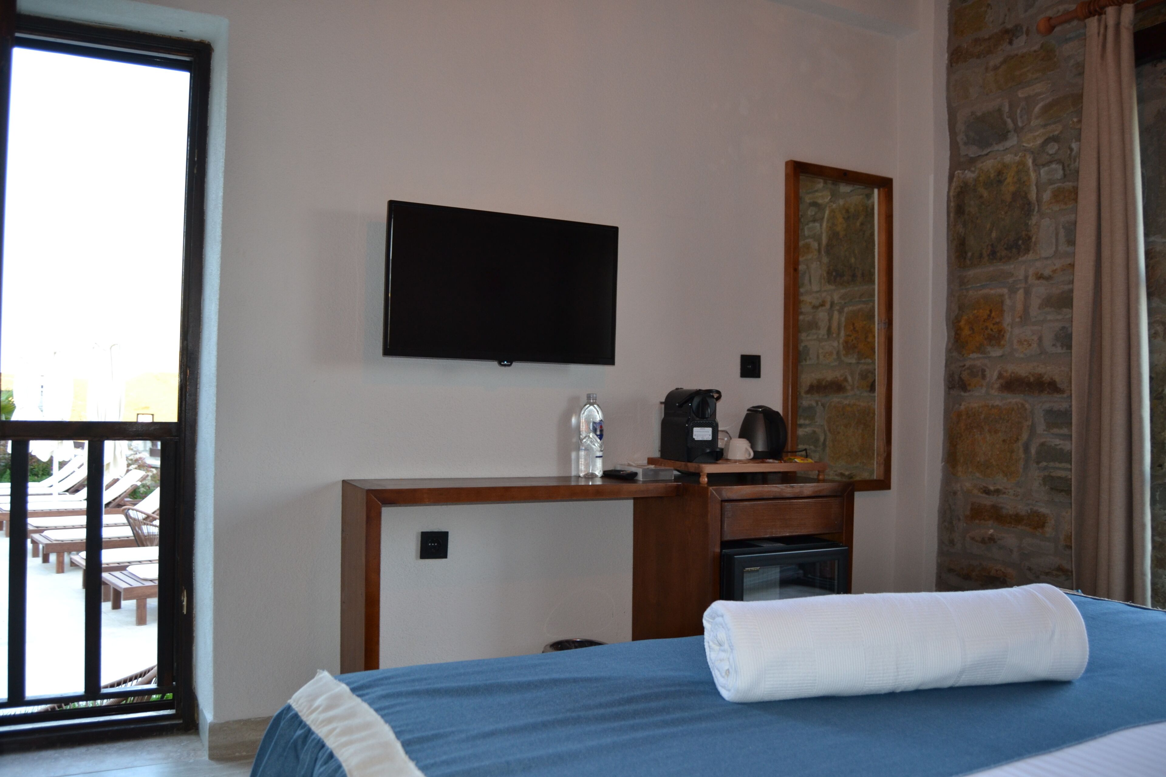 Une chambre d’hôtel avec un lit, un téléviseur à écran plat fixé au mur, un bureau avec une machine à café et un mur en pierre.