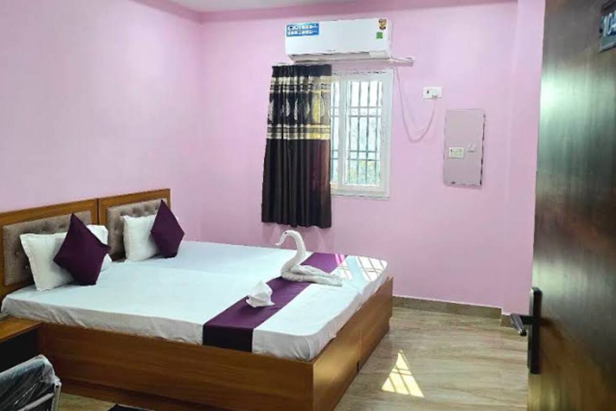 A hotel room with a bed, a ceiling fan, an air conditioning unit, a window with curtains, and a door.