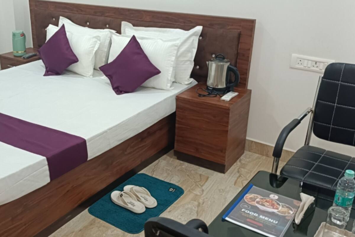 A neatly made bed with a purple accent, a wooden headboard, a nightstand with a kettle, a pair of slippers, and a table with a food menu.