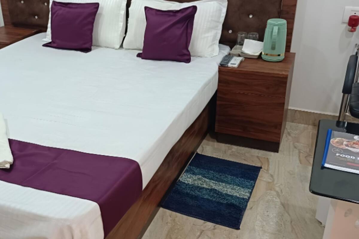A neatly made bed with a white and purple bedspread, a wooden headboard, a nightstand with a green water bottle, and a blue rug on the floor.