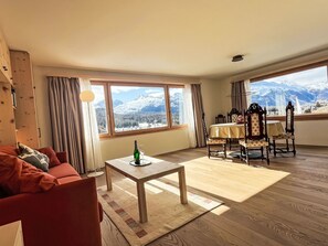Een woonkamer met een bank, een tafel met een fles en een glas, een eethoek met stoelen en een groot raam met uitzicht op de bergen.
