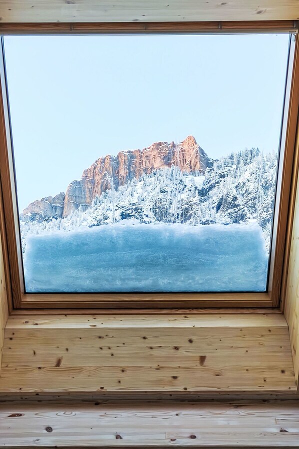 A wooden-framed window with a view of a snow-covered mountain and forest.