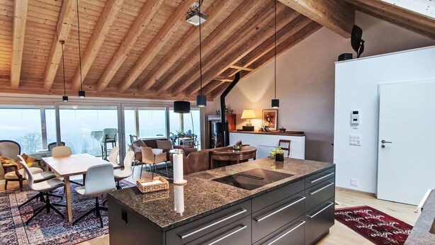 Een moderne keuken met een groot schiereiland, houten balken in het plafond en uitzicht door grote ramen.