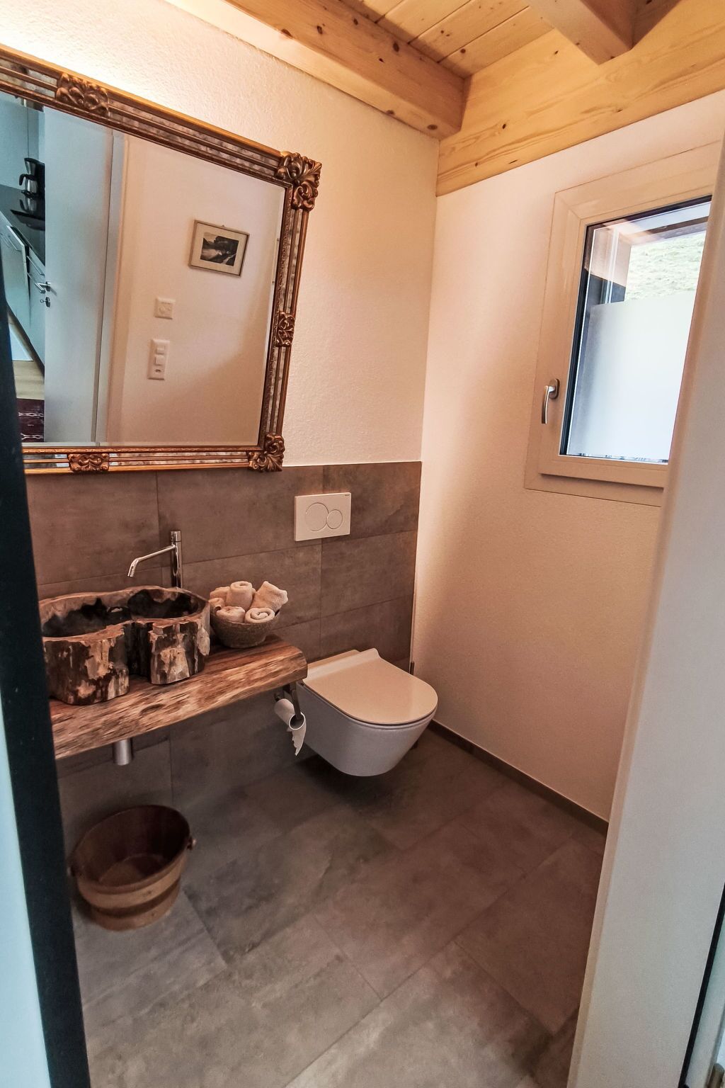 A bathroom with a wooden vanity, a toilet, a mirror with an ornate frame, and a window.